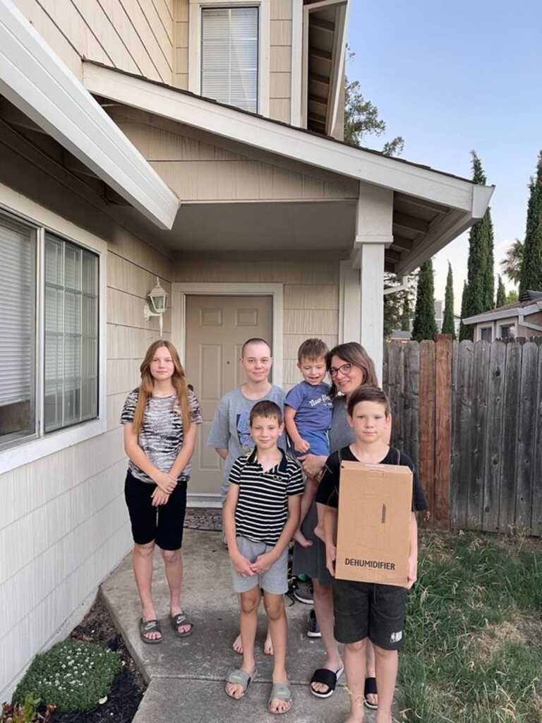 A family of four smiles while standing in their front yard on a sunny day.