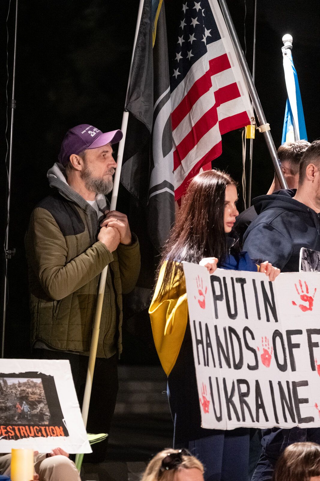 A man holds a flagpole beside the American and Ukrainian flags while a woman next to him holds a "Putin hands off Ukraine" sign.