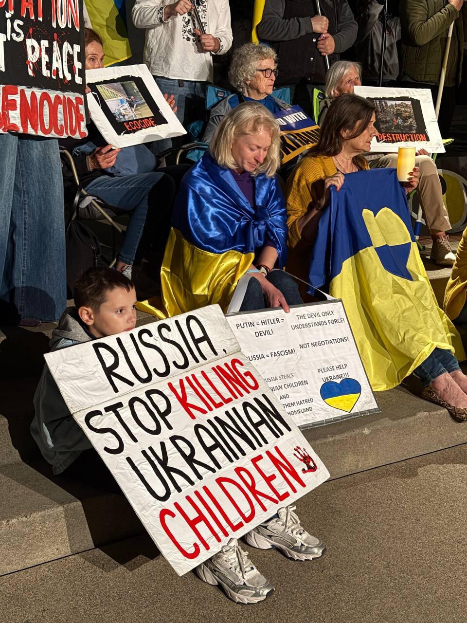 A boy sits on the Capitol steps holding a sign that reads "Russia, stop killing Ukrainian children", next to women holding Ukrainian flags.